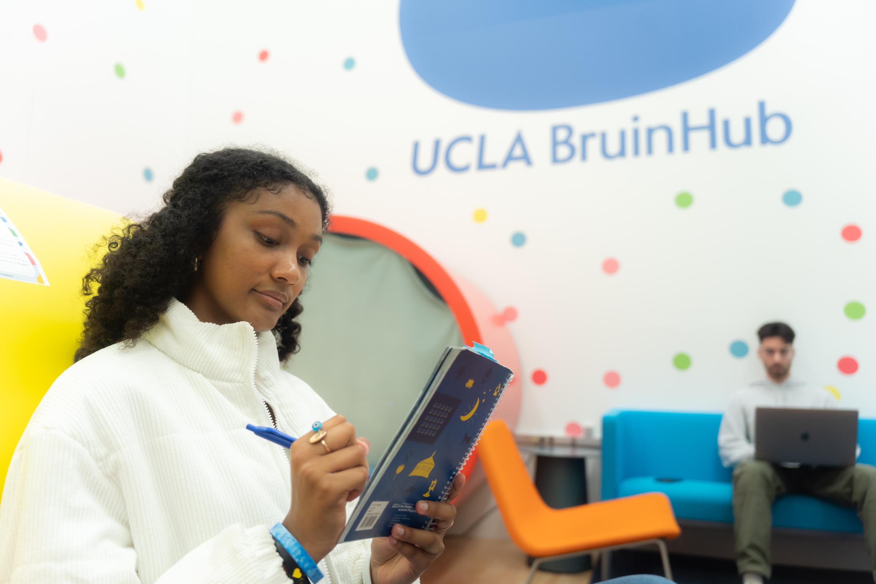 Photo of a student writing in their notebook, sitting in front of a BruinHub pod in the JWC. Another student is seen in the background on their laptop, sitting in the JWC lounge